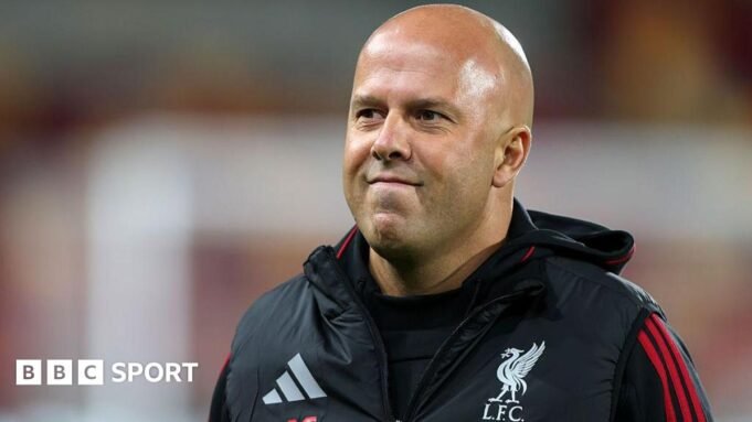 Arne Slot smiles and is wearing a black coat with a Liverpool FC logo on the chest
