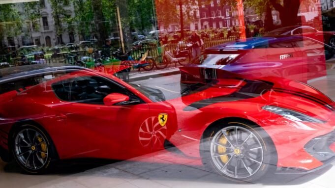法拉利削減英國交付量 Red Ferrari cars on display in a showroom, with reflections of trees and people outside visible in the glass.